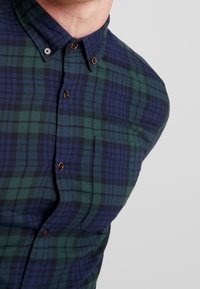 Camicia button-up a quadri verdi e blu, con colletto, bottoni marroni, tasca sul petto sinistro e tessuto testurizzato.