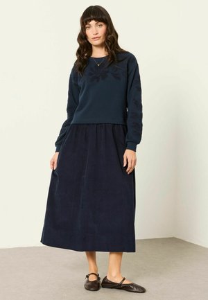 Donna con capelli scuri e mossi indossa una maglia ricamata blu navy a maniche lunghe, gonna midi coordinata e ballerine nere Mary Jane, in piedi al chiuso.