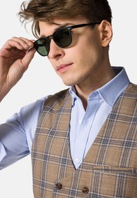 Brązowa kamizelka w kratę z niebieskimi akcentami na jasnoniebieskiej koszuli; czarne okulary przeciwsłoneczne z ciemnozielonymi soczewkami, trzymane jedną ręką blisko twarzy.