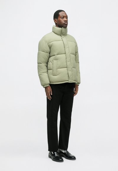 Oliwkowa kurtka puchowa z wysokim kołnierzem i pikowanym wzorem, z dwoma przednimi kieszeniami. Noszona z czarnymi spodniami i butami.