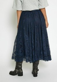 Gonna midi in pizzo blu navy con pieghe e modelli floreali intricati, rifinita con un orlo scallop, abbinata a stivali neri in pelle al ginocchio.