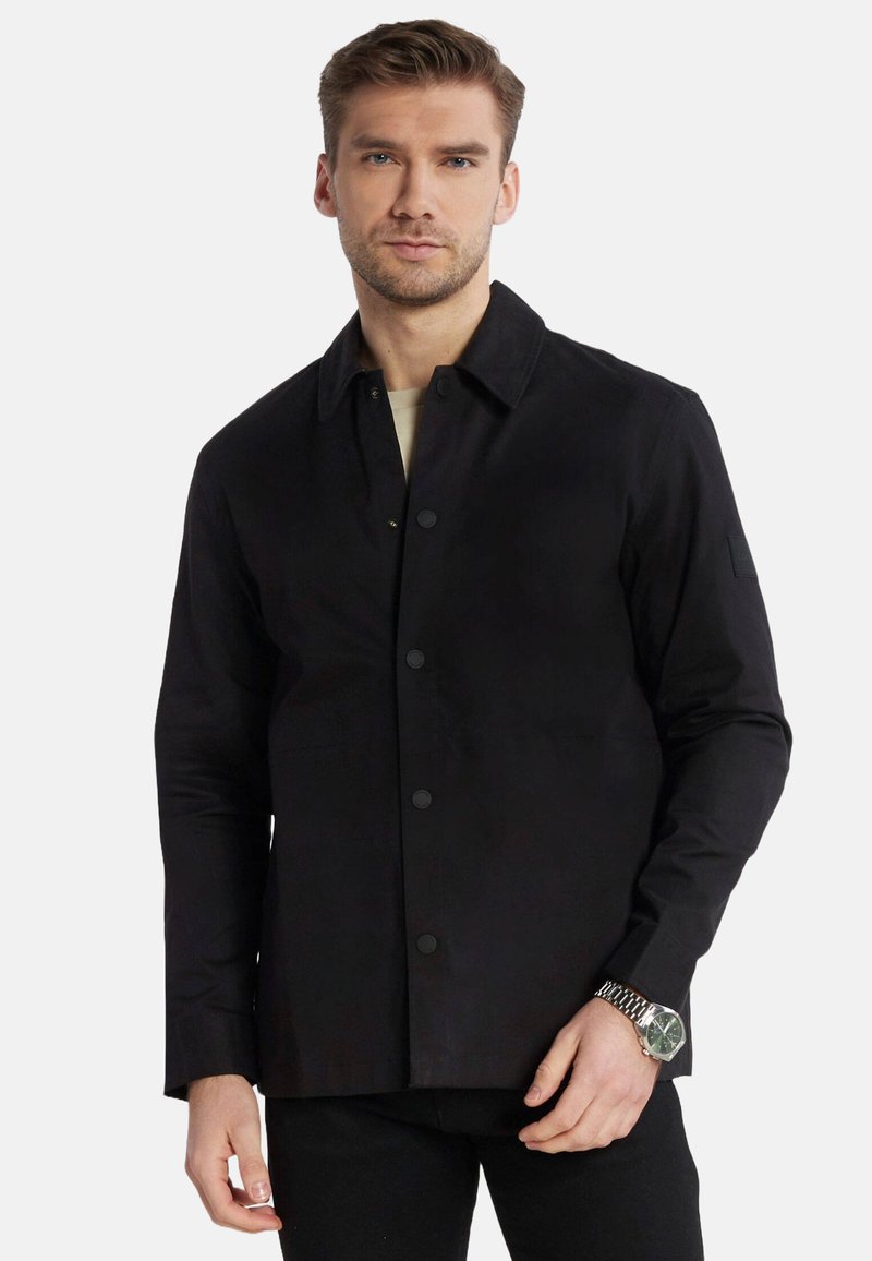 Giacca nera con chiusura a bottoni, maniche lunghe e colletto. Presenta una texture liscia e include un logo discreto sul braccio sinistro.