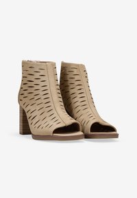 Botines de tobillo abiertos en color beige, hechos de suave ante, con patrones recortados, cierre de cremallera y un tacón de bloque texturizado que imita la madera.