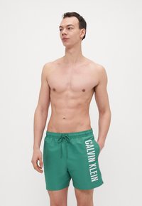 Grønne svømmeshorts laget av glatt materiale med elastisk midjebånd og snøring. Har hvit "CALVIN KLEIN"-merkevare på siden.
