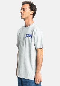 Gray cotton t-shirt with short sleeves, featuring a blue "D S H O E S" logo on the left chest. The fit is relaxed, paired with blue denim jeans.