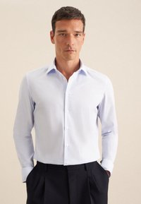 Camicia azzurra chiaro a maniche lunghe con sottili righe verticali, dotata di colletto classico e taschino, abbinata a pantaloni scuri.