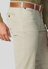 Beige katoenen broeken met een rechte pijp, een achterzak met knoopsluiting en een bruine leren riem met metalen gesp.