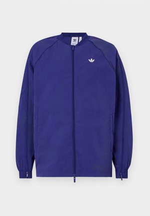 Violeta viegla jaka ar rāvējslēdzēju, elastīgām aprocēm un mazāku baltu logotipu uz krūtīm. Gluda tekstūra un ikdienas dizains.