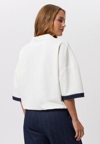 Sweat-shirt blanc surdimensionné avec un col, présentant des poignets en denim bleu rayé. Texture lisse et coupe décontractée mises en évidence dans le design.