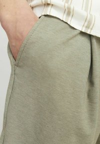 Pantalons en mélange de coton vert clair avec détail de poche latérale, présentant une texture lisse et une coupe décontractée, partiellement couverts par un tissu rayé.