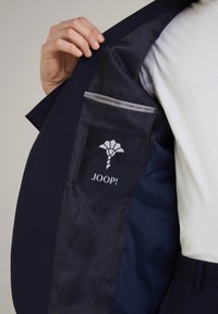 Wnętrze granatowego żakietu cechuje gładka satynowa podszewka z powtarzającym się wzorem, kieszeń na piersi z logo JOOP! oraz szary akcent.