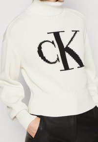 Pull à col roulé blanc côtelé avec logo "CK" noir. Comprend des manches longues et un ourlet ajusté. Fabriqué en maille texturée.