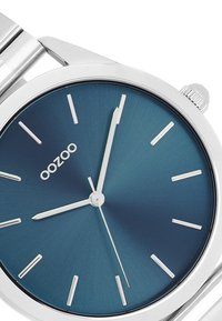 Montre en métal argenté avec un boîtier rond, un cadran dégradé bleu, des index et aiguilles argentés, présentant une surface texturée et une couronne sur le côté.