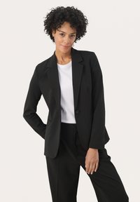 Blazer noir en tissu lisse, avec une fermeture à un bouton, des revers à cran et des poches latérales, porté sur une chemise blanche.