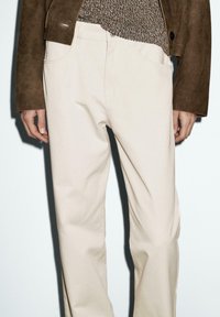 Beige straight-leg pants worn with a brown suede jacket and a brown knit sweater, showing hands resting by the sides.