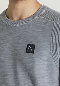 Maglione grigio a maglia con scollatura a coste e pannello spessorato sulle spalle. Un piccolo logo bianco su fondo nero è posizionato sul petto.