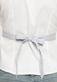 Λευκή μπλούζα με κορδόνι στο πίσω μέρος από ριγωτό ύφασμα. Το κορδόνι διαθέτει λεπτομέρεια φιόγκου. Ομαλή υφή και προσαρμοσμένη εφαρμογή εμφανείς.
