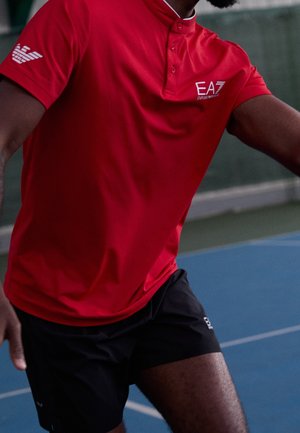 Mężczyzna w czerwonym polo EA7 Emporio Armani i czarnych szortach gra na niebieskim zewnętrznym boisku sportowym.