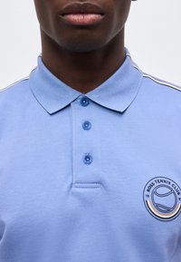 Άνδρας που φοράει ανοιχτό μπλε πόλο με τρία κουμπιά και το λογότυπο "Boss Tennis Club" στο αριστερό στήθος.