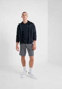 Námořnický větrovka, tričko s krátkým rukávem tmavé barvy, šedé a námořnické pruhované šortky, bílé tenisky, postávající v minimalistickém vnitřním prostředí.