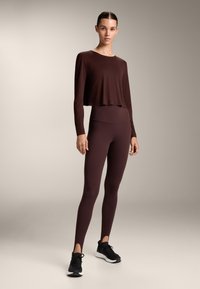 Nainen seisoo, päällään tummanruskea pitkähihaisen cropped-toppi ja siihen sopivat korkeavyötäröiset leggingsit mustilla urheilukengillä.