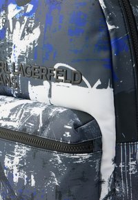 Σακίδιο πλάτης με γκρι ύφασμα που διαθέτει σχέδιο με πιτσιλιές μπογιάς σε μπλε και λευκό. Περιλαμβάνει μια μεγάλη τσέπη με φερμουάρ και ανάγλυφη λεπτομέρεια με λογότυπο.