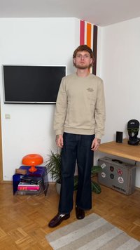 Felpa beige con logo, jeans in denim scuro e scarpe marroni lucide. Lo sfondo presenta una TV, strisce colorate e piante da interno.