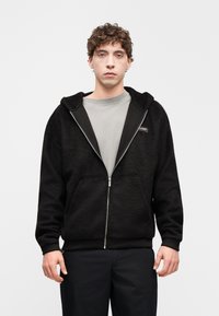 Sweat à capuche zippé noir en tissu texturé avec une finition douce, comprenant une capuche, des poches avant, et un t-shirt gris col rond en dessous.