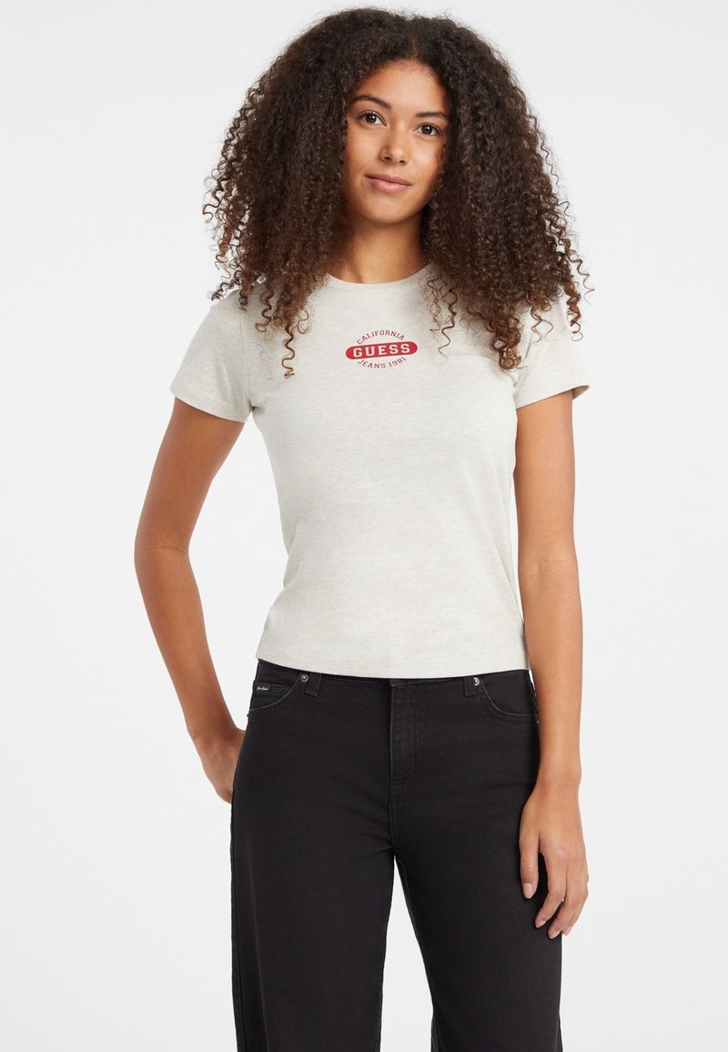 Hellgraues T-Shirt mit kurzen Ärmeln, featuring ein rotes "Guess"-Logo und Textdesign. Kombiniert mit schwarzen Jeans, die eine figurbetonte Silhouette zeigen.