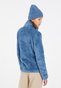 Giacca in pile blu con una finitura morbida e testurizzata e colletto alto, abbinata a un berretto coordinato con un motivo intrecciato.