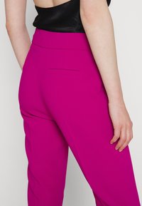 Vrouw in felroze hoge taillebroek met een paspelzak aan de achterkant en een zwarte mouwloze top.