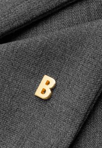 Szary materiał o gładkiej teksturze z wyraźną złotą metalową szpilką w kształcie litery "B". Projekt jest prosty i minimalistyczny.