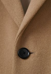 Țesătură de palton din lână maro cu o textură moale, dotată cu o guler în formă de lăcaș și un buton negru cu patru găuri care accentuează designul.