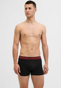 Schwarze Boxershorts mit einem roten Logo-Bund, glatte Textur, enganliegendes Design und Länge bis zur Mitte des Oberschenkels. Keine sichtbaren Muster oder Verzierungen.
