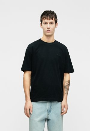 T-shirt de algodão preto com mangas curtas, decote redondo e um discreto logotipo no peito, combinado com jeans de algodão azul claro.