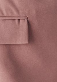 Gestructureerde stof in een zachte rozenkleur met een opvallend gevouwen pocketdetail. Het oppervlak heeft een subtiel diagonaal geweven patroon.