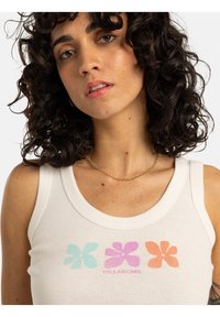 Débardeur blanc côtelé avec trois graphiques de fleurs colorées : bleu clair, rose et orange, avec le mot "BILLABONG" en violet.