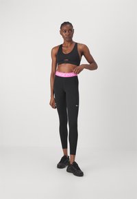 Černá sportovní podprsenka s výřezovým designem, kombinovaná s černými legínami s růžovým páskem "NIKE PRO", obuté s černými sportovními botami.