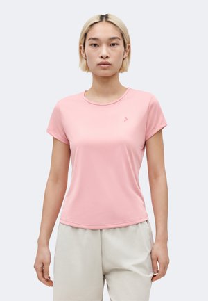 Young person with short blonde hair wearing a fitted light pink short-sleeve shirt and light beige pants, standing straight facing forward.