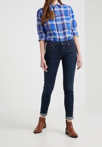 Blå rutiga knapp-up skjorta, mörka slim fit jeans, bruna ankelskängor. Upprullade ärmslut på jeansen, skjortan har en avslappnad passform och strukturerat mönster.