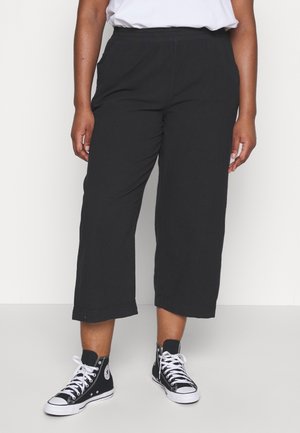 Personne portant un pantalon large noir raccourci, un t-shirt blanc et des baskets montantes noires avec des lacets blancs, debout sur un sol blanc.