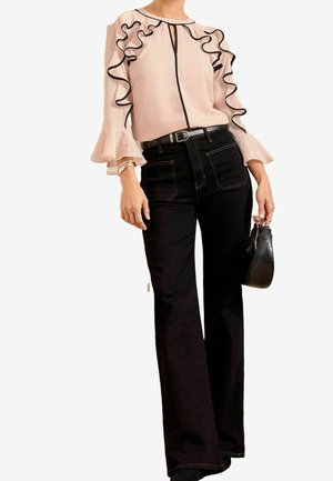 Kobieta w jasnoróżowej bluzce z czarnym falbankowym obszyciem i rozszerzanymi rękawami, czarne spodnie z wysokim stanem, trzymająca czarną teksturowaną torebkę.