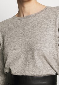 Gros plan sur une personne portant un pull en tricot gris clair rentré dans une jupe en cuir noire taille haute, montrant le cou et le haut du torse.