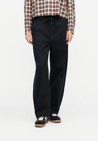 Άτομο που φοράει μαύρο παντελόνι φαρδύ με κορδόνι στη μέση, κόκκινο και άσπρο πουκάμισο καρό, και μαύρα αθλητικά παπούτσια με λευκά κορδόνια.