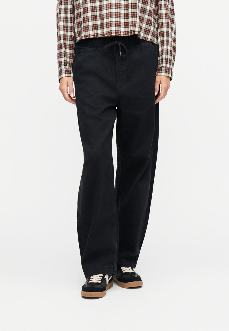 Άτομο που φοράει μαύρο παντελόνι φαρδύ με κορδόνι στη μέση, κόκκινο και άσπρο πουκάμισο καρό, και μαύρα αθλητικά παπούτσια με λευκά κορδόνια.