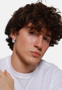 Silberne spiralige Ohrringe, runde Form, mit polierter Textur. Die Person trägt ein weißes T-Shirt, hat lockiges dunkles Haar und einen neutralen Hintergrund.