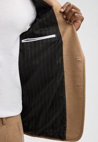 Brauner Wollblazer mit glattem Innenfutter, das schwarze und goldene vertikale Streifen aufweist. Enthält eine diskrete Innentasche mit einem weißen Reißverschluss.