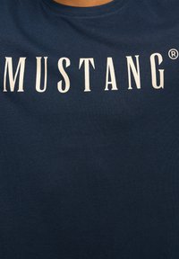 Tamsiai mėlynas medvilninis marškinėlis, ant kurio krūtinės yra didelis baltas užrašas "MUSTANG®". Trumpomis rankovėmis ir apvaliu kaklu.