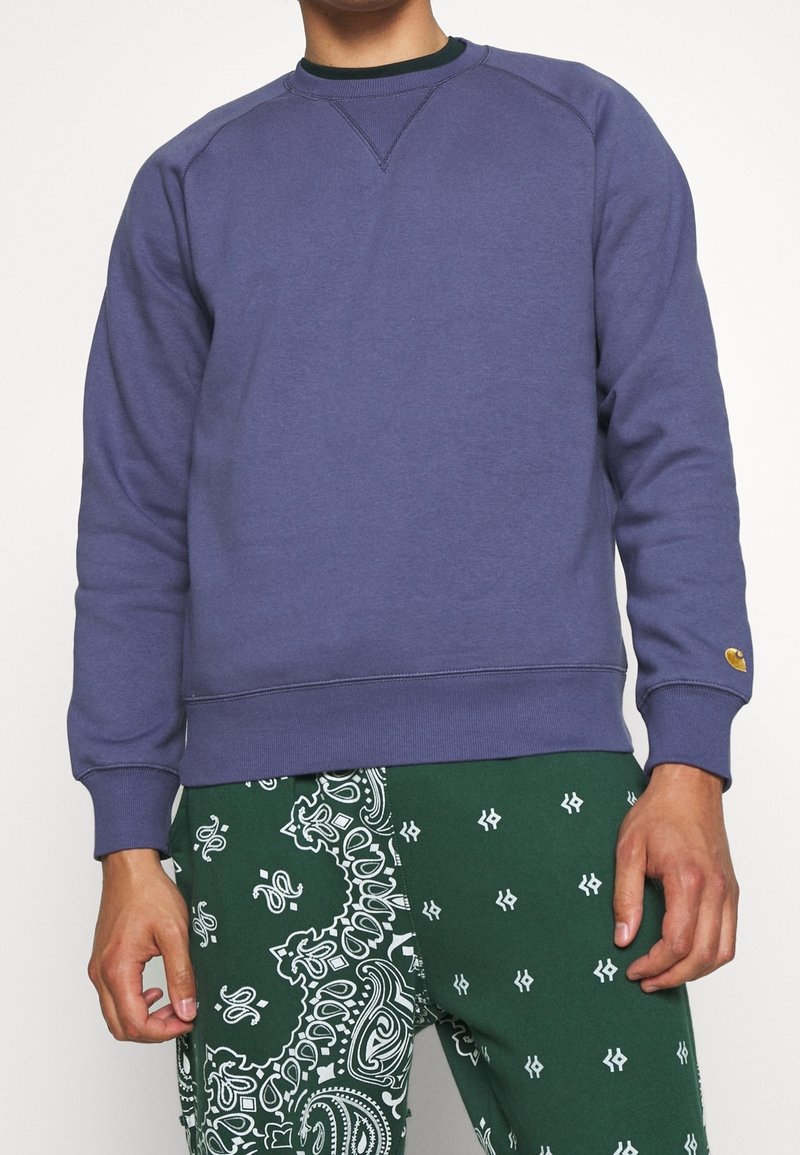 Sudadera de algodón morada con mangas largas, escote redondo y puños acanalados. Exhibida con pantalones verdes que presentan un patrón paisley.