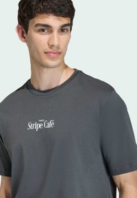 Šedé tričko z měkké látky s kulatým výstřihem a bílým potiskem "Three Stripe Café" na hrudi. Jednoduchý, neformální design.